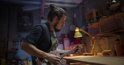 Young handsome Hispanic guy carpenter checking handcraft design musical instrument indoors, pleasant satisfied male master luthier holding guitar feeling happy about result of work with wood