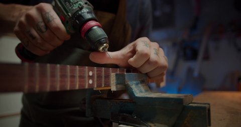 Close up of handsome powerful joiner male artisan drilling thin screw in wooden guitar, work process of confident young Hispanic guy making musical instrument using drill tool for wooden planks