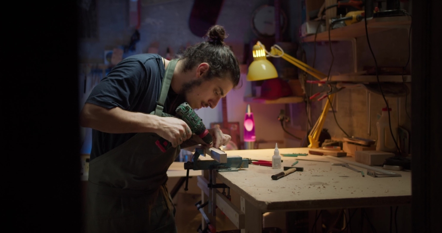 Side shot of attractive brunette guy drilling wooden guitar neck musical instrument, professional male young artisan joiner in dark light working on wood table in cozy workshop, art and hobby concept | Shutterstock HD Video #1057516705
