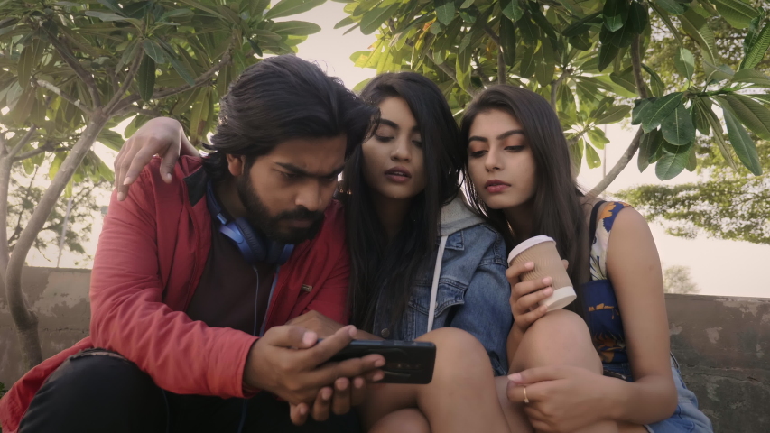 A group of young male and female friends are sitting outdoors and watching a live sport using mobile phone or smartphone. Fashionable and happy millennial celebrating when favourite team scores.  Royalty-Free Stock Footage #1057516864