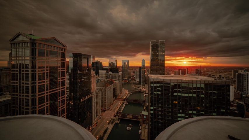 Zoom in aerial time lapse of downtown Chicago with boats on river passing bridges along Wacker Drive as sun sets below the horizon lighting up clouds and lights come on in the city at night.