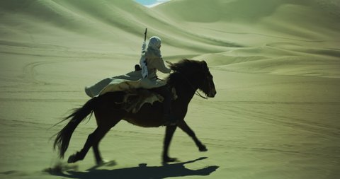 Masked person rides a galloping horse through the desert. Sand dunes surround a person riding a horse with a rifle on their back. Long journey. Cinematic, Shot on RED