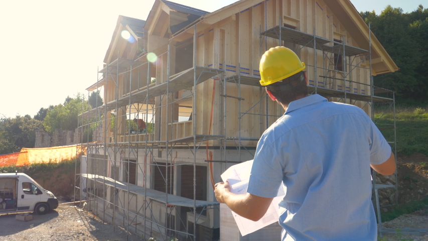 SLOW MOTION, CLOSE UP, LENS FLARE: Unrecognizable construction site manager looks at the floor plans while standing outside of a modern CLT house under construction in the countryside on a sunny day. Royalty-Free Stock Footage #1057523074