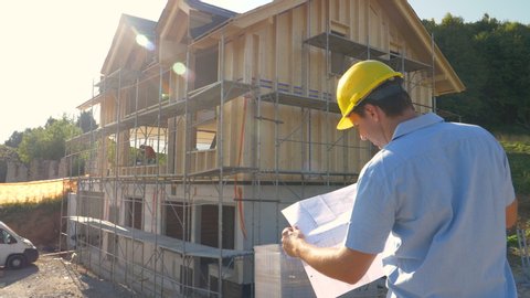 SLOW MOTION, CLOSE UP, LENS FLARE: Unrecognizable construction site manager looks at the floor plans while standing outside of a modern CLT house under construction in the countryside on a sunny day.