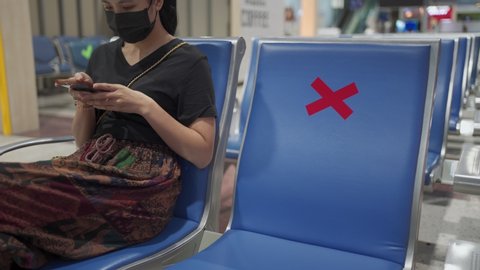 Asian woman wear black mask sit down on chair bench, social distance concept check sign cross sign, 2 meters distancing, new normal During COVID-19 Pandemic. Inside airport terminal, using cellphone