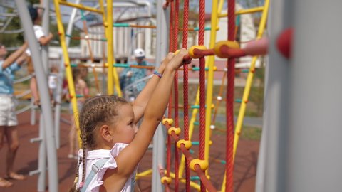 Little girl with pigtails and dressed in denim overalls climbs rope wall or ladder on playground. Happy child climbing up on playground net, active lifestyle