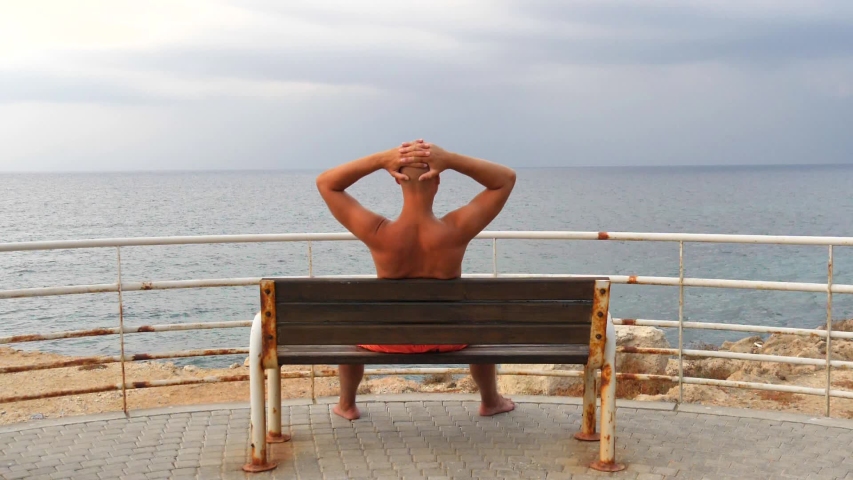 A young bald tanned man with a naked torso sits on an bench with his back, starters to sea. Сoncept of rest, relax, vacation alone, contemplation, wanderlust. Slow motion video. Island of Cyprus.