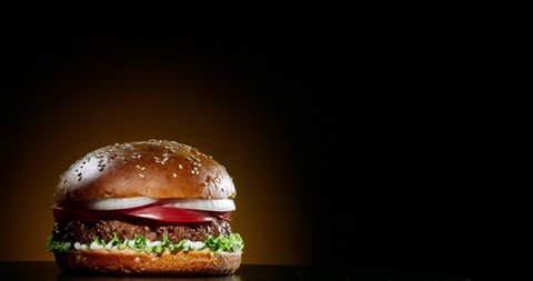 Big appetizing burger is rotating on the black background with copy space. Delicious hamburger with fresh iceberg cabbage, onions, tomatoes and grilled steak. Rotating - fast food, 4k footage.