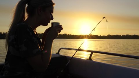 A woman is drinking tea from a thermos while fishing. Fishing on the lake from a boat. Fishing rods in sunlight. Fishing rest concept.