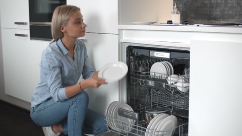 Young beautiful woman unloading dishes from dishwasher machine. Pretty female taking white plates from dishwasher in kitchen. Easy housework with kitchen appliances concept
