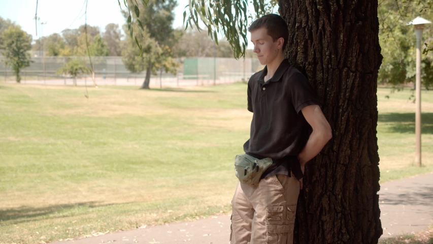 Man Leans Back Against Tree Standing In Shade, Sunny Day With Open Grassy Field In Background