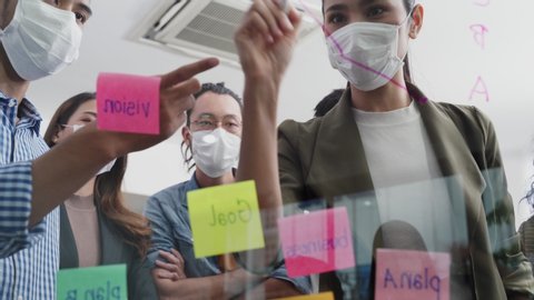 Group brainstorming of Asian people putting sticky note, put it on window. Teamwork business people wearing mask prevent covid infection having discussion and share an idea to solve problem in office.
