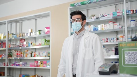Middle eastern male pharmacist wearing protective hygienic mask to prevent infection selling medications to woman patient and making drug recommendations in modern pharmacy