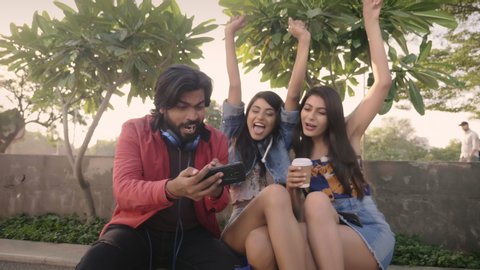 A group of young male and female friends are sitting outdoors and watching a live sport using mobile phone or smartphone. Fashionable and happy millennial celebrating when favourite team scores. 