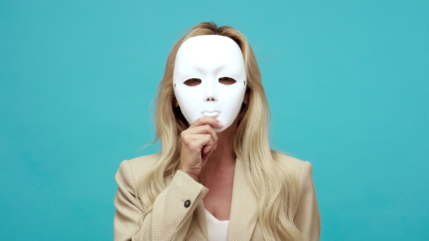 Confident middle aged woman with blond hair in business style jacket putting off mask showing different emotions, duplicity, hypocrisy. Indoor studio shot isolated on blue background Royalty-Free Stock Footage #1057545973