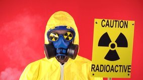 Man in Protective Chemical Suit, Protective Mask with a Poster Caution Radiation. Turns A Finger At The Temple. Studio video, Behind a man smoke, glasses with radiation signs, on a red background.