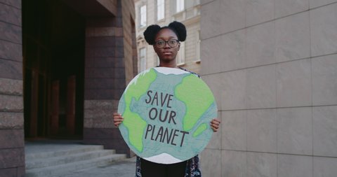Afro american girl with carton placard looking to camera while supporting eco movement. Millennial woman holding earth model with save our planet writing while standing at street.