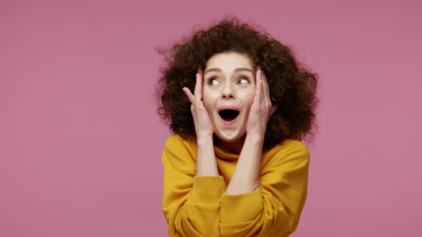 Oh my god wow! Amazed excited  young woman afro hairstyle in hoodie raising hands in surprise looking at camera with big eyes, shocked by sudden victory. indoor studio shot isolated on pink background