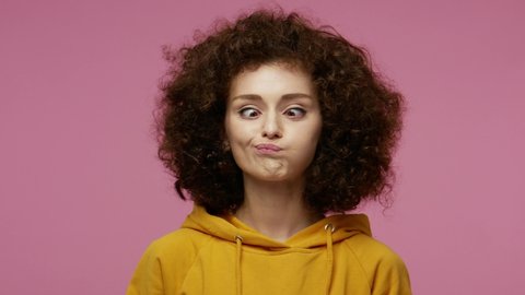 Funny amusing young funny woman in hoodie looking awkward with crossed eyes and puffed cheeks, fooling around making stupid brainless dumb expression. indoor studio shot isolated on pink background