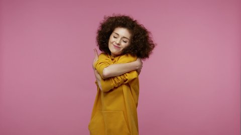 Adorable young woman in afro hairstyle in hoodie embracing herself, hugging with self-love and smiling from pleasure, narcissistic egoistic feelings, complacency concept. isolated on pink background