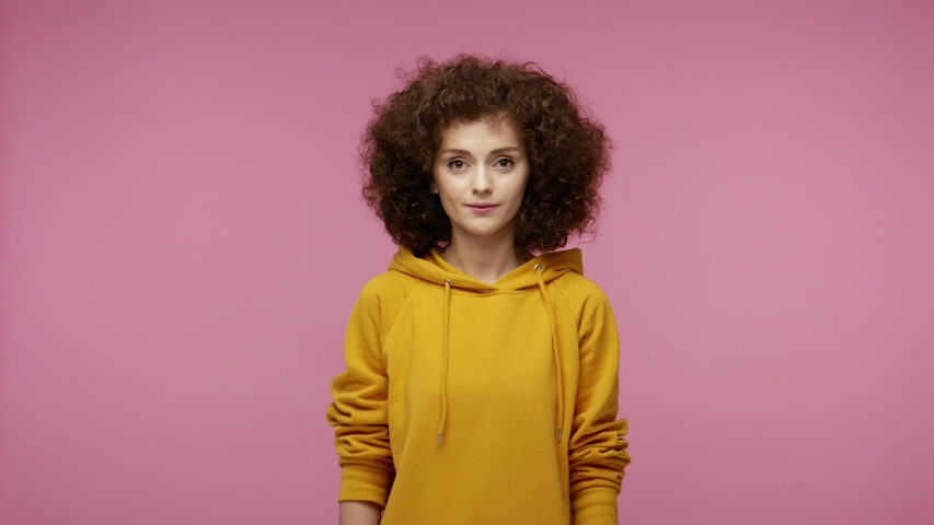 Sociable happy  young woman afro hairstyle in hoodie smiling friendly at camera and waving hands gesturing hello or goodbye, welcoming with hospitable expression. indoor isolated on pink background Royalty-Free Stock Footage #1057547146