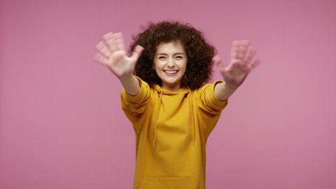 Sociable happy  young woman afro hairstyle in hoodie smiling friendly at camera and waving hands gesturing hello or goodbye, welcoming with hospitable expression. indoor isolated on pink background