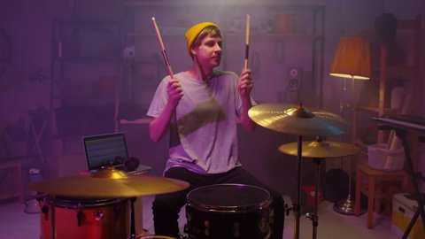 Slowmo shot of cheerful young adult Caucasian guy wearing casual outfit with yellow knit cap enjoying playing drums in his garage studio