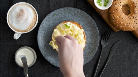 Bagel with cream cheese and scrambled eggs. Female hands prepare healthy breakfast sandwich with toasted bagel, cream cheese, scrambled eggs and scallions