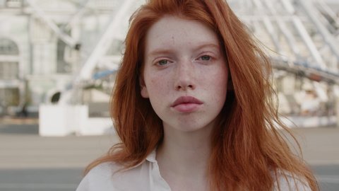 Attractive red-haired young woman with cute face freckles looking serious into camera outside the street city center. Beautiful ginger. Female portraits.