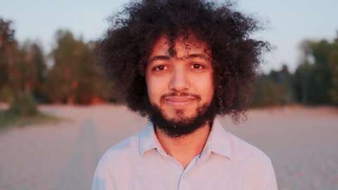 Close-up Portrait of Joyful Young Hispanic Man Curly Hair, Standing Alone in Park, Smiling and Looking at Camera. Stylish Authentic People, Expressive Charismatic Independent Person.