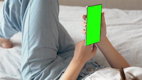Young woman relaxing on her bed with her mobile phone zooming out from the blank screen in an over the shoulder view