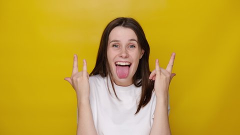 Happy cute young girl makes rock n roll sign, yells loudly, look at camera, dressed in white t-shirt, devil horns hand sign gesture, stands over yellow studio background, feels self confident