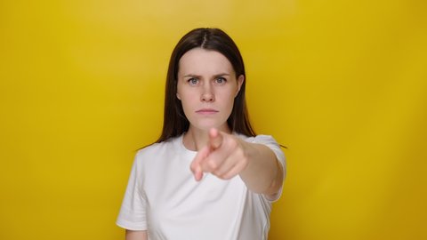 Angry brunette girl pointing finger to camera, looking dissatisfied, frowning and accusing with strict negative expression, choosing guilty, wears white t-shirt, isolated on yellow studio background