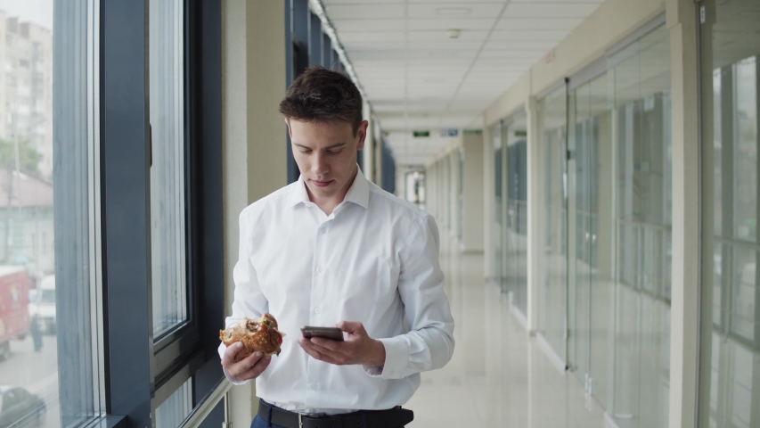 Handsome businessman in suit types on the mobile phone, eats the burger and looks out the window with smile in light corridor of office centre. 4K