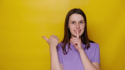 Young woman point with finger at blank copy space aside, girl make hush be quiet gesture, tell about show secret sale offer, dressed in purple t-shirt, isolated on yellow studio background