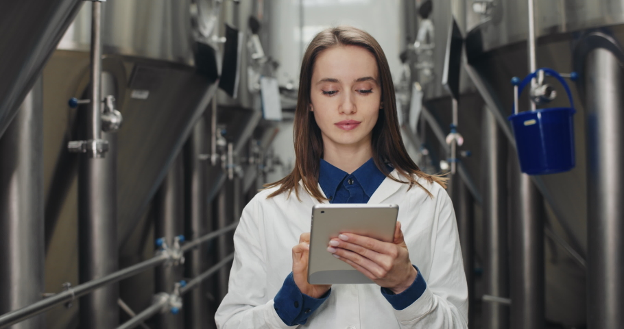 Handsome female process engineer maintaining records on tablet at brewery. Woman worker touching and typing digital display while walking and looking at on equipment gauges. Royalty-Free Stock Footage #1057570258