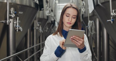 Handsome female process engineer maintaining records on tablet at brewery. Woman worker touching and typing digital display while walking and looking at on equipment gauges.