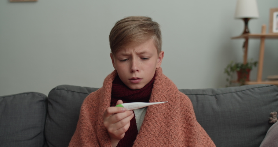 Ill boy having cold and measuring temperature while sitting on sofa. Teen kid covered in blanket looking at thermometer and shaking head. Concept of illness and healthcare. | Shutterstock HD Video #1057570282