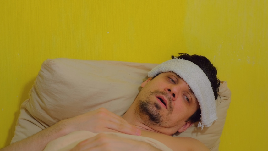 A tired sick man waking up with headache, fever, sars, cold. lying in bed covering blanket, portrait closeup. Rising in temperature. Coronavirus pandemic, epidemic. Yellow background.