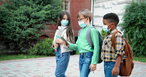 Multi-ethnic junior students in masks with backpacks going to school. Caucasian girl with copybooks. African American boy with schoolbag. Little kid watching something on smartphone. Study concept