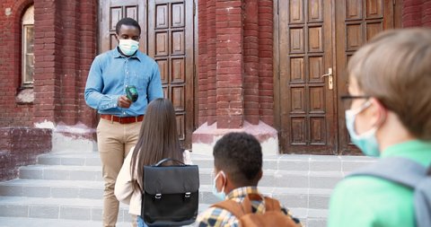 African American handsome male teacher measuring temperature of multi-ethnic pupils with infrared thermometer in front of school. After quarantine study. Healthcare control Preventive measures concept