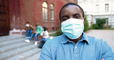 Close up portrait of African American handsome man in mask looking away and smiling to camera near school. Male teacher outdoor. Mixed-races junior students on background. Quarantine concept