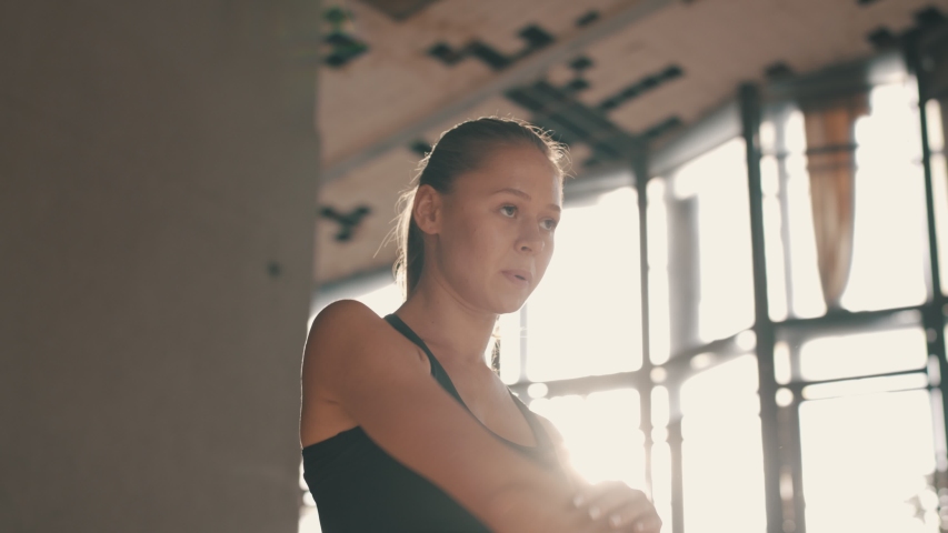 Confident Sporty Young Woman in Black Sportswear Stretching Arms and Back Muscles before Workout. Athletic Woman Training for Competition in Sunlit Abandoned Factory Building. Slow Motion.