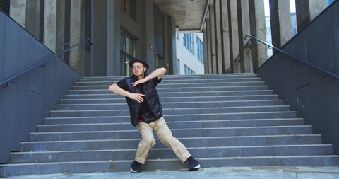 Happy handsome Caucasian guy performs modern hip hop dance on street in city. Young joyful man dancing modern choreography on stairs outdoors in good mood. Freestyle moves. Street dancing concept