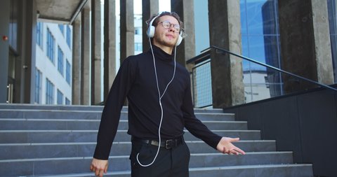 Portrait of happy young man listening to music in headphones and dancing on stairs outdoor in town. Handsome Caucasian joyful male in glasses listens to songs and moving on street. Leisure concept