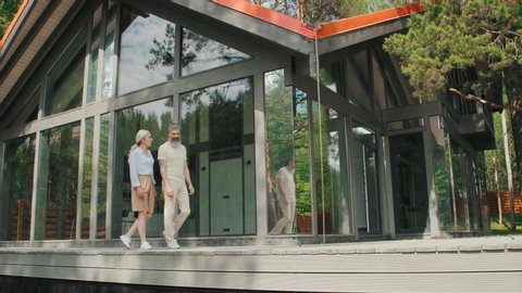 Tracking shot of happy upper-class senior couple walking along porch of modern house with panoramic windows and chatting