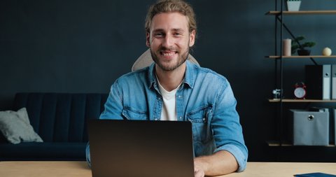 Young Successful Freelancer is sitting at Home Office, smiling at the Camera. Portrait of Satisfied Young Man is working on his Laptop at Home, looking directly in Shoot. Casual Style. Freelance.