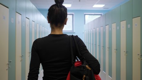 slim woman with bun hairstyle in black clothes walks carrying sports bag along empty brightly lit gym dressing room slow motion backside view