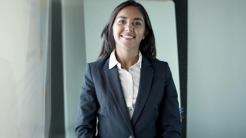 Portrait of Latin pretty young businesswoman standing in office room. Happy brunette senior female manager in suit with folded arms smiling and looking at camera. Human resources and personnel concept