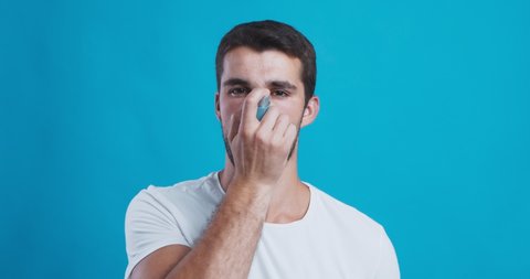 Young man suffering from asthma attack using breathing inhaler, blue studio background